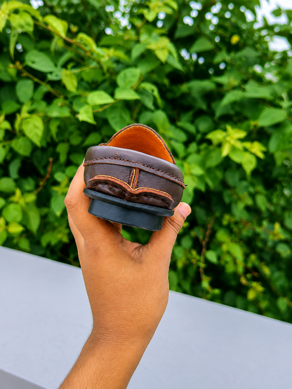 Back view of men's soft leather shoes held in hand against a green leafy background.